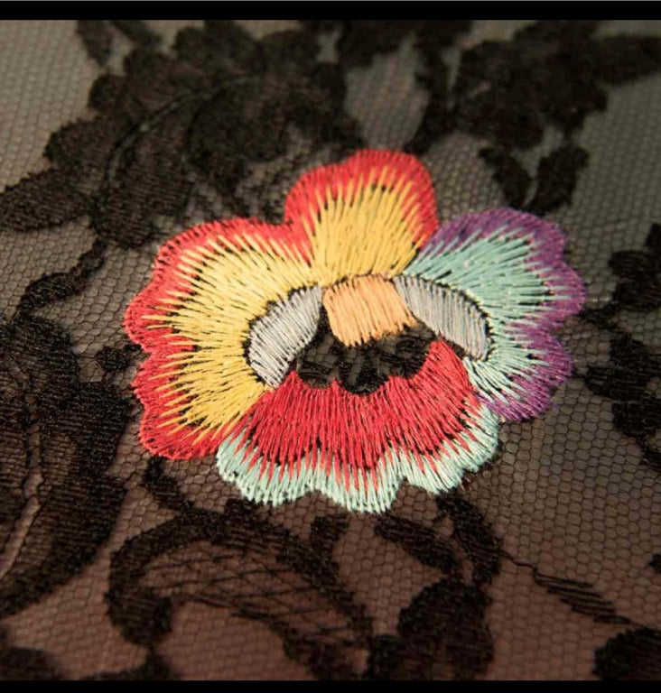 Close up of a colourful hand embroidered flower on a black lace against white background.