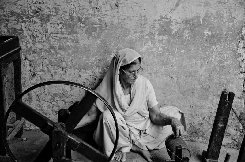 An old Indian woman handspinning yarn for Ezma while sitting on the ground against a distressed wall.